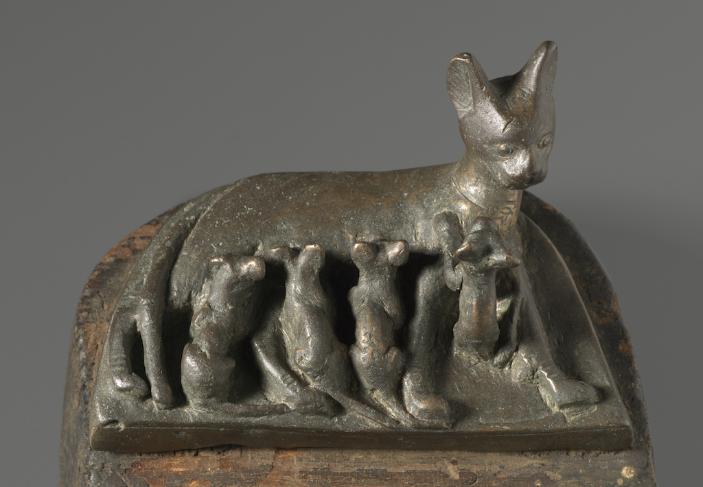 Bronze group of a mother cat with four kittens on a bronze base (A) set into a wooden base (B). The cat reclines upon her left side with her legs outstretched. Her left fore and hind legs lie forward while her right fore and hind legs are drawn back. Her tail is draped over her right hind leg. Her head is upright perhaps tilted back slightly as one of the kittens, set between her forelegs, playfully climbs toward her neck. The next three kittens are being nursed by the mother cat. Two are between her fore and hind legs, the last is between the cat's tail and left foreleg. Mother cat wears a necklace which is indicated by an incised line running around her neck. A pendant? Perhaps a wadjet(?) is incised in the front of the necklace. The five felines are upon a semi-circular base with straight front and curved back. A pair of tangs below the bronze base fit into the original wooden base, also semi-circular. The wooden base was originally painted black. On the front of the bronze base toward the right side (below the cat's left paw) is an inscription Bastet,